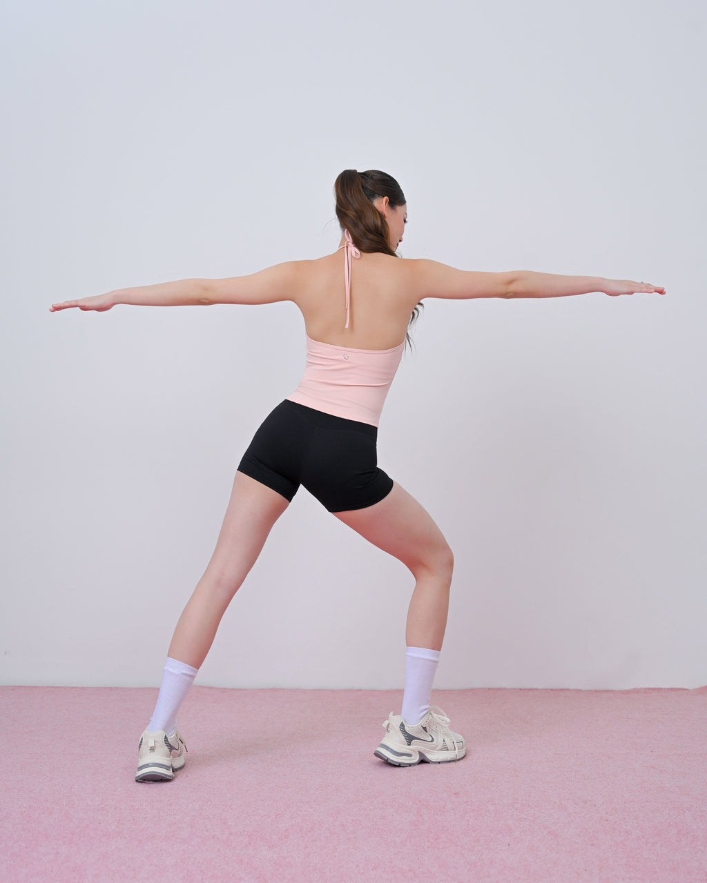 A woman in a light pink sports bra and black athletic shorts poses in a yoga warrior II stance, viewed from behind, against a minimalist light gray wall and pink floor.