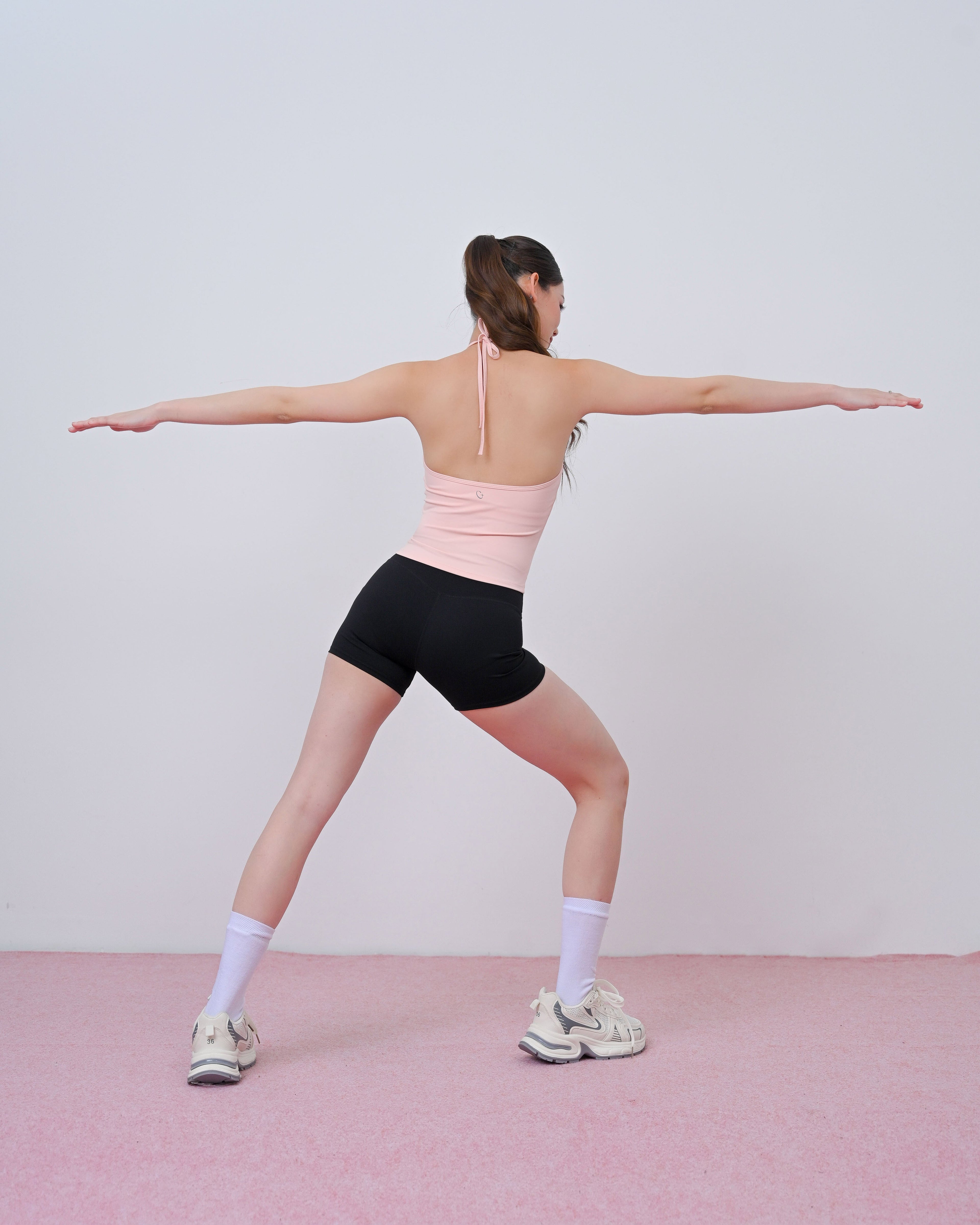 A woman in a light pink sports bra and black athletic shorts poses in a yoga warrior II stance, viewed from behind, against a minimalist light gray wall and pink floor.